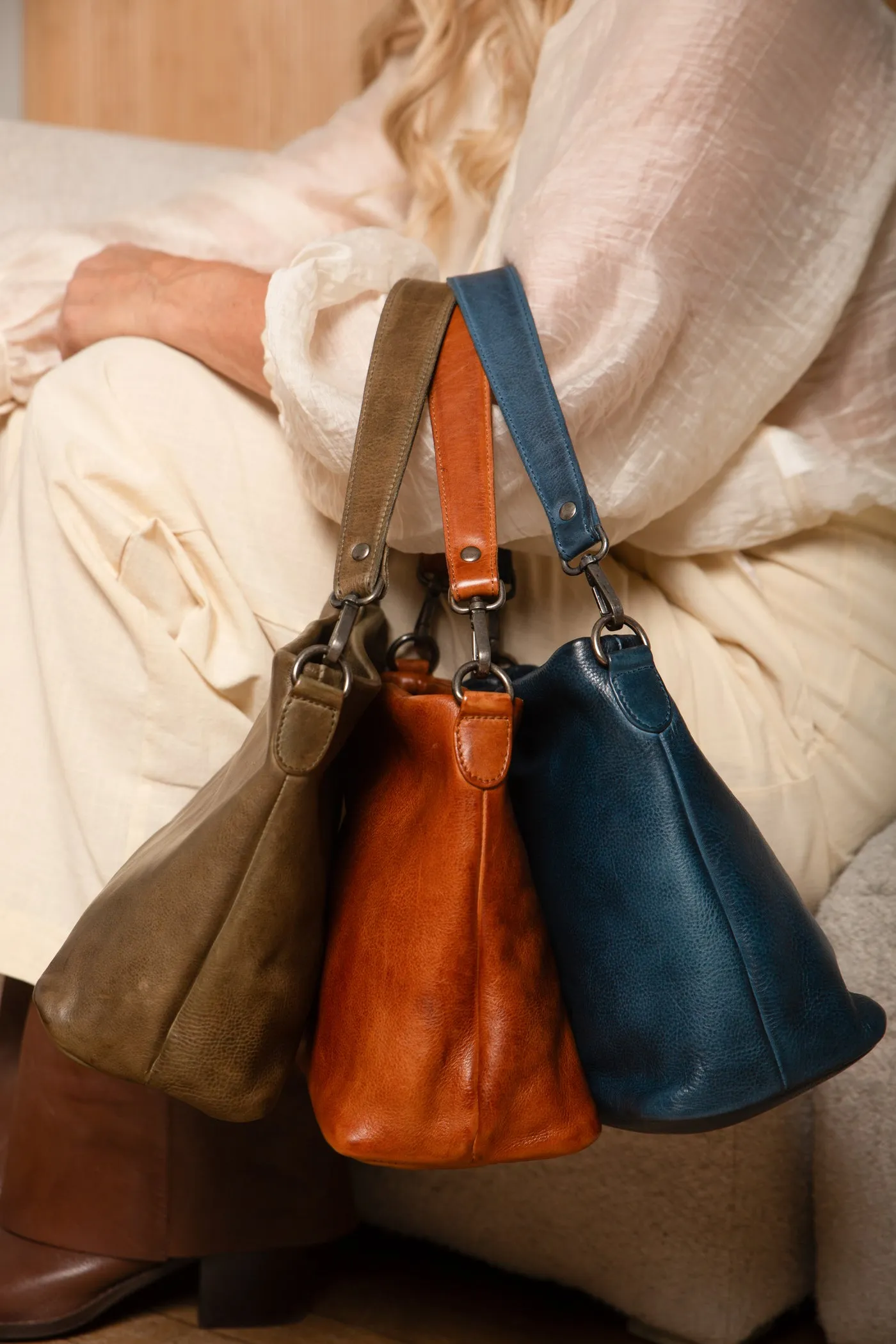 A woman holds Latico Ivy crossbody and shoulder bags in moss, cognac and indigo, from the Latico about page.